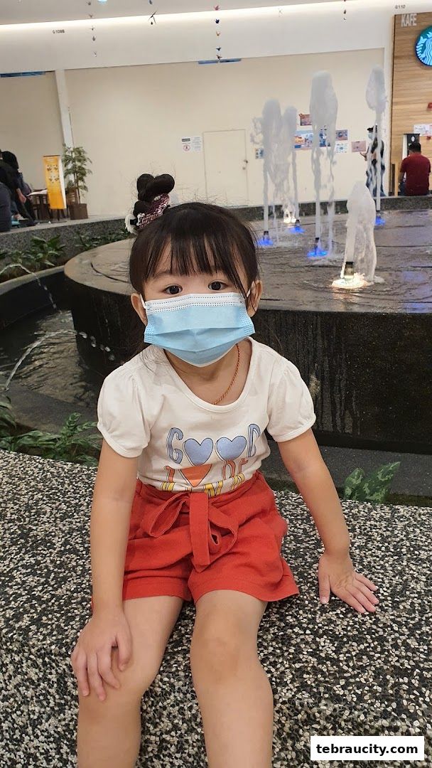 A young girl wearing a mask sits near a fountain in a shopping mall.