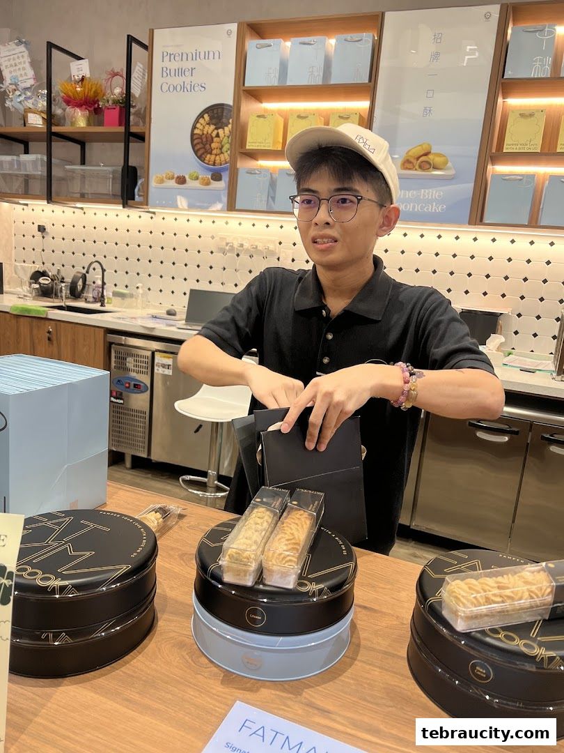 A young man stands behind a counter displaying Fatmama cookies in decorative tins.