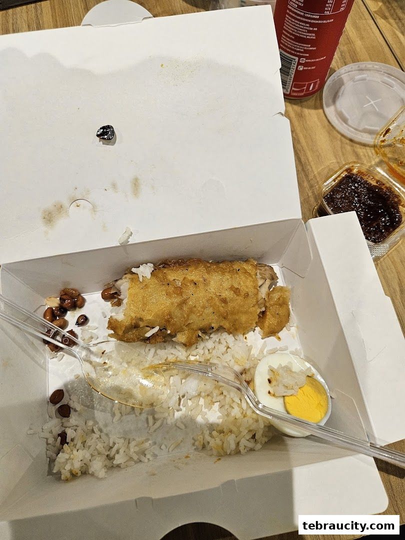 A takeaway box filled with rice, fried chicken, peanuts, and a hard-boiled egg, with chili sauce and a drink nearby.