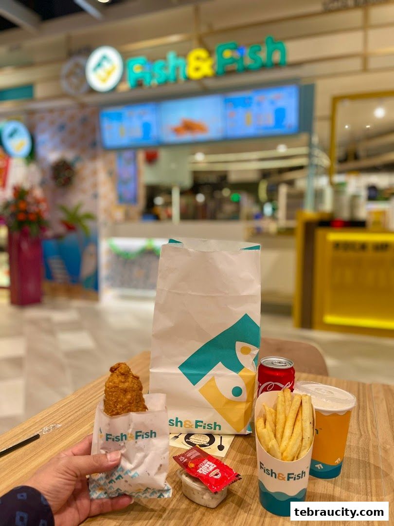 A person holds a piece of fried fish from Fish & Fish, with fries, a drink, and a soda on the table.