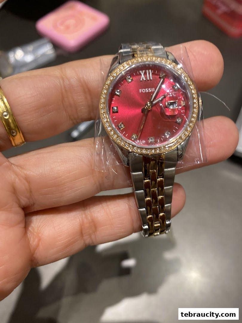A hand holds a Fossil watch with a red dial and a two-tone metal band.