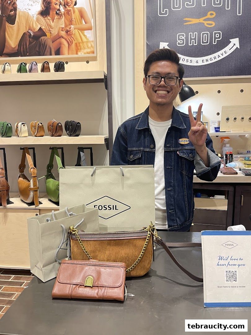 A smiling employee stands behind a counter with Fossil bags and wallets.