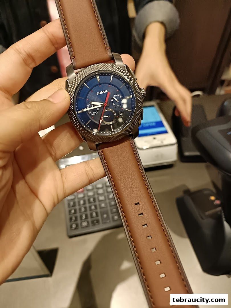 A hand holds a Fossil watch with a brown leather strap in front of a store counter.