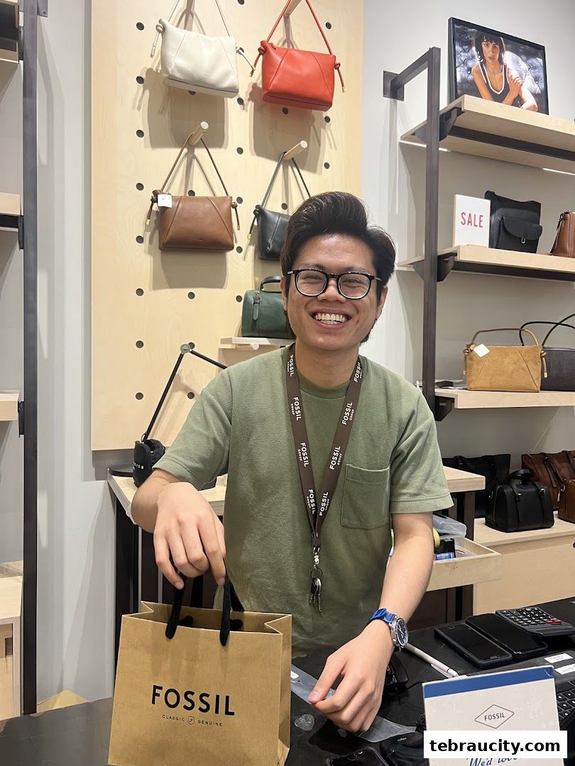 A smiling employee holds a Fossil shopping bag in a store displaying various handbags.