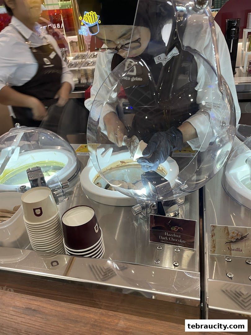 A gelato shop employee serves a customer, with various gelato flavors displayed.