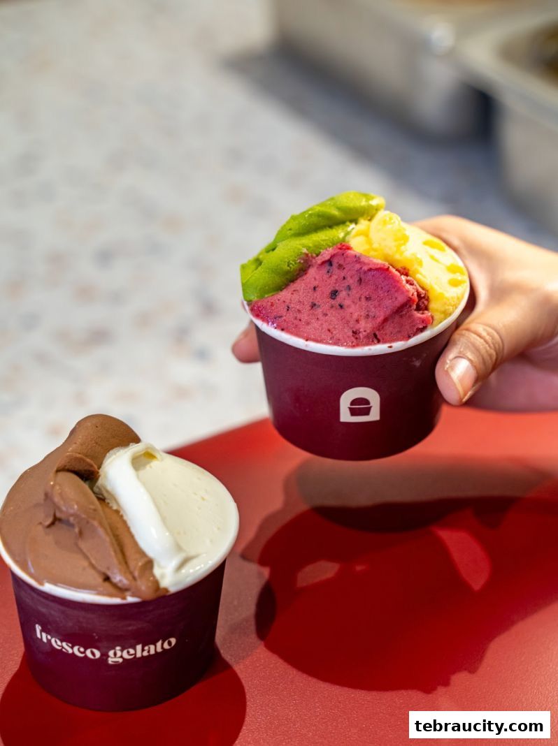 A hand holds a cup of colorful gelato, with another cup of gelato in the foreground.