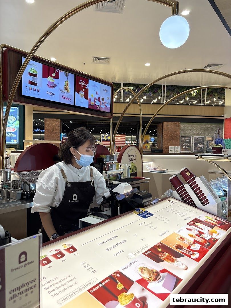 A staff member serves gelato at a Fresco Gelato counter with digital menus and product displays.