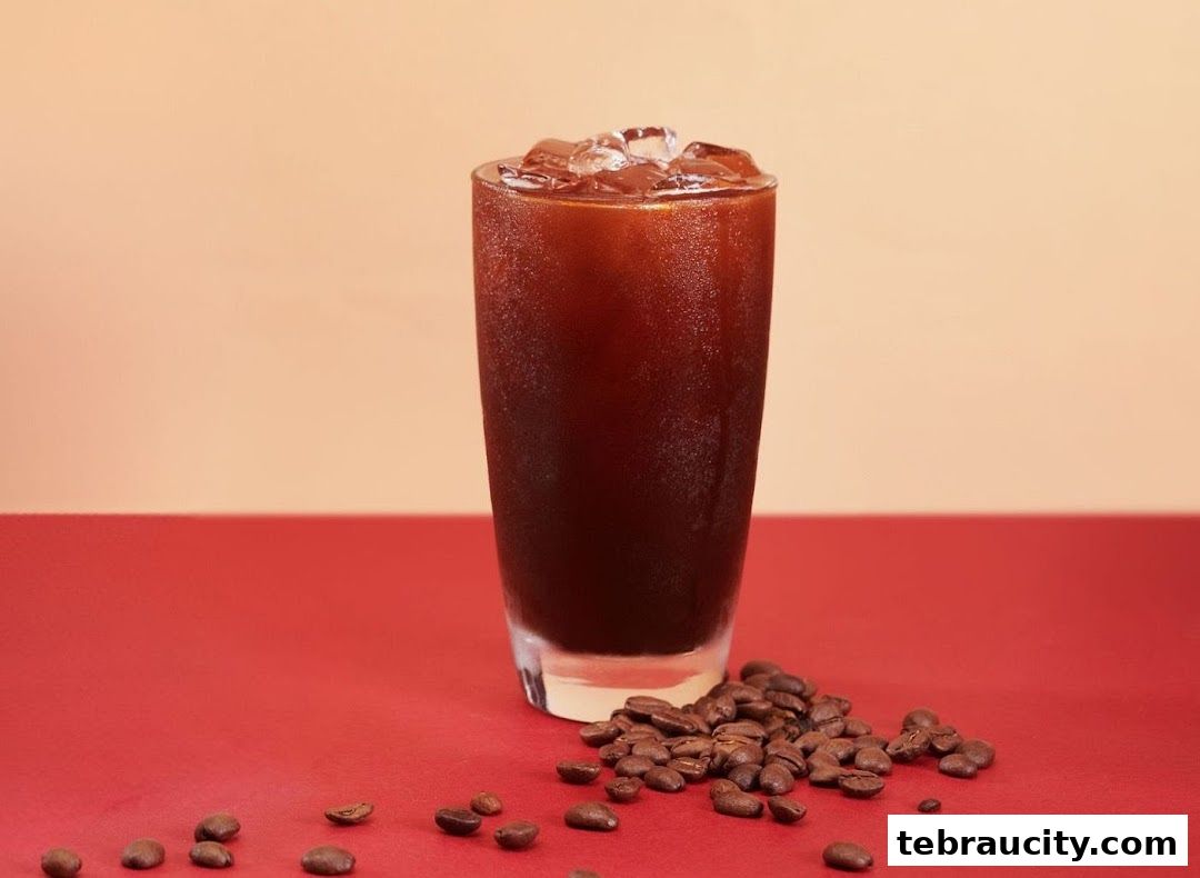 A tall glass of iced coffee with coffee beans scattered around it.