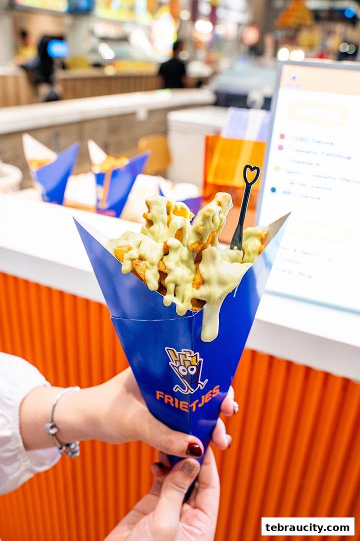 A person holds a cone of waffle fries generously topped with a creamy green sauce.