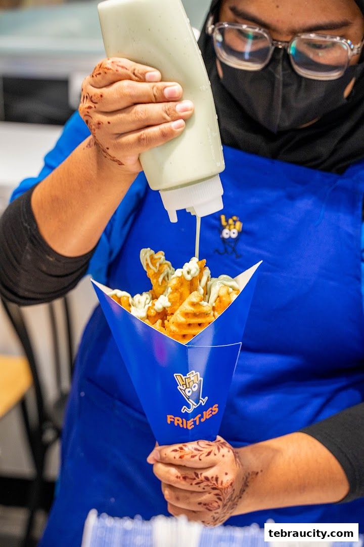 A person is drizzling sauce on a cone of waffle fries with the Frietjes logo.