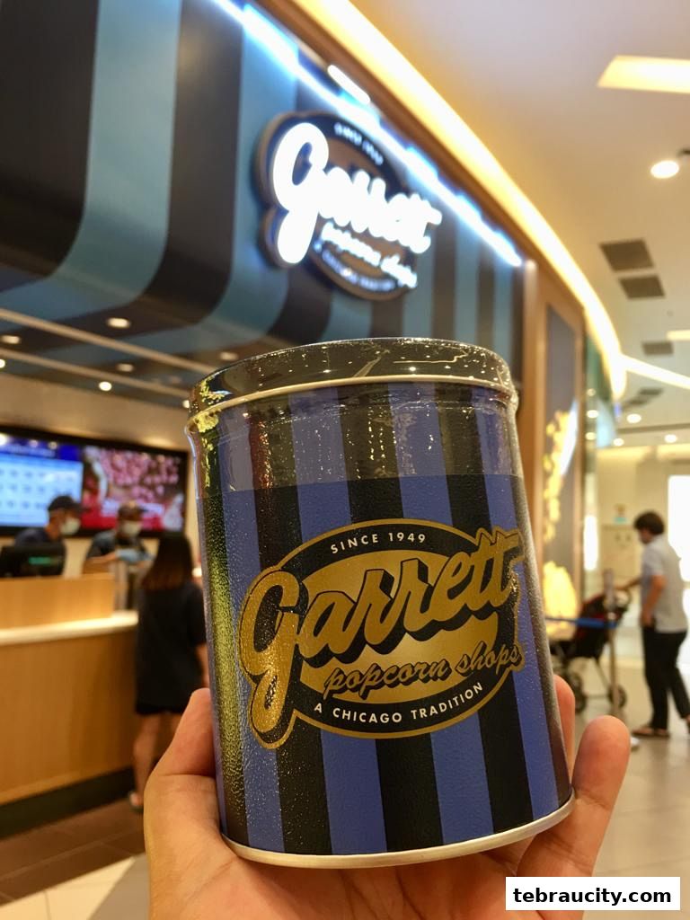 A hand holds a tin of Garrett Popcorn in front of the shop's entrance.