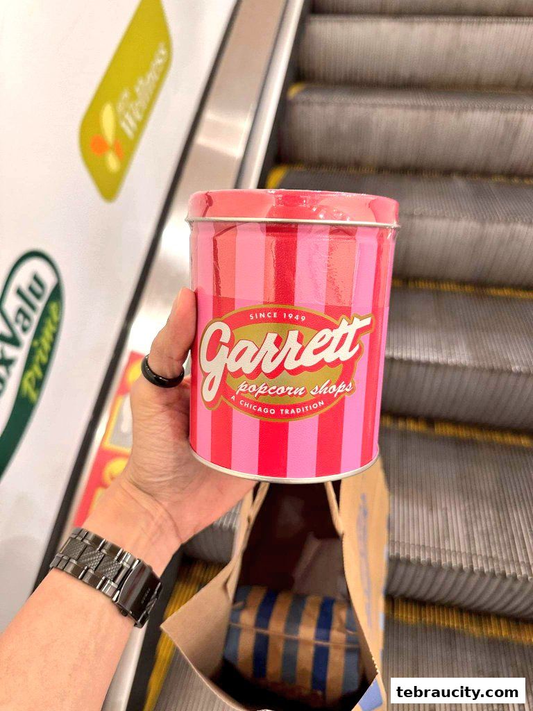 A hand holds a pink and red striped Garrett Popcorn Shops tin near an escalator.