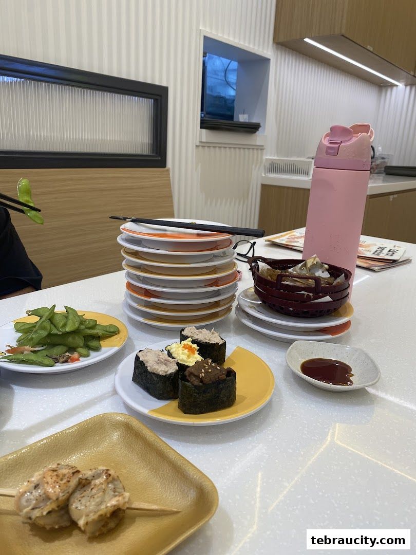 A table at Genki Sushi with various sushi dishes, edamame, and a stack of plates.