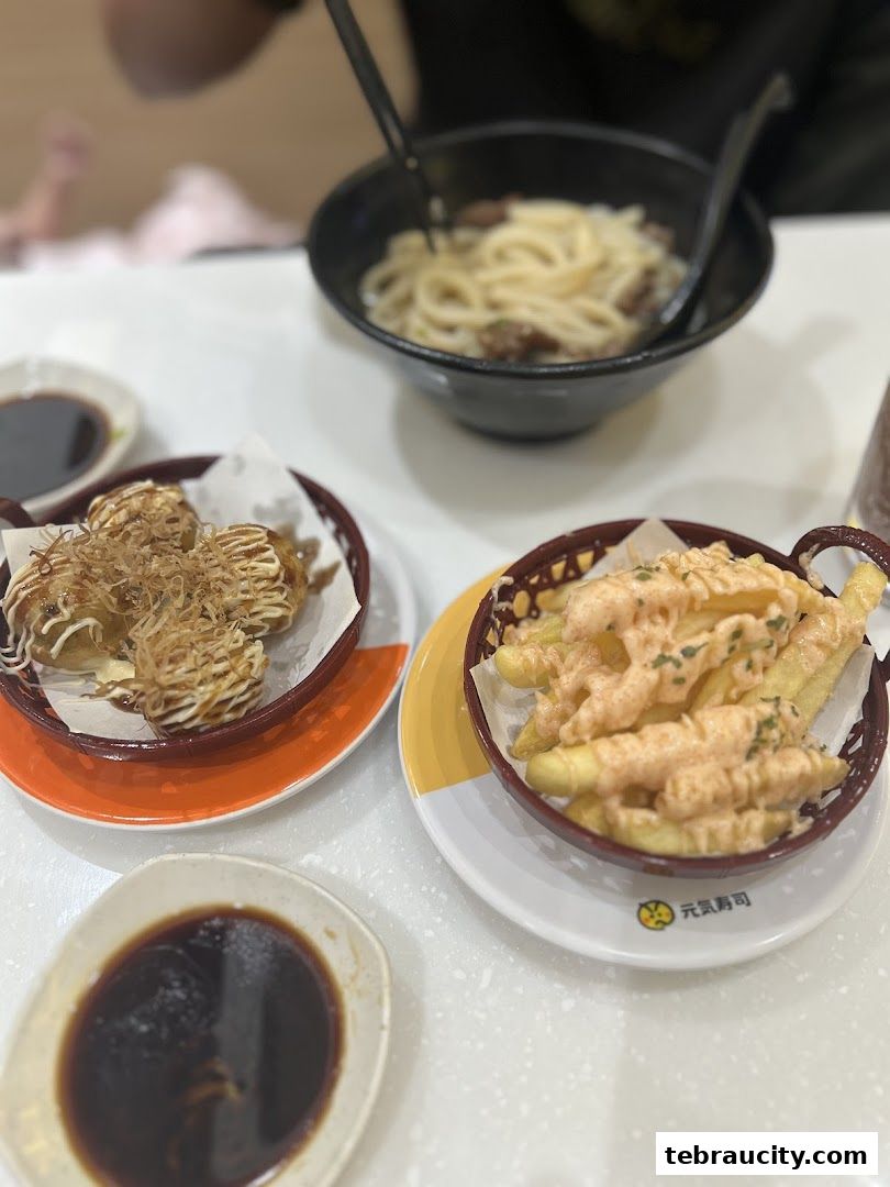 A table at Genki Sushi with various Japanese dishes including takoyaki, fries, and udon noodles.