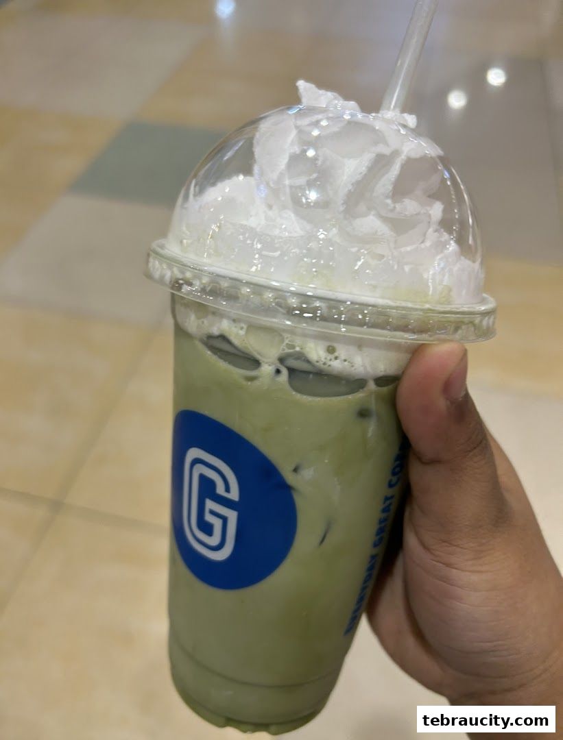 A hand holds a tall, clear cup filled with a green beverage and topped with whipped cream and ice.