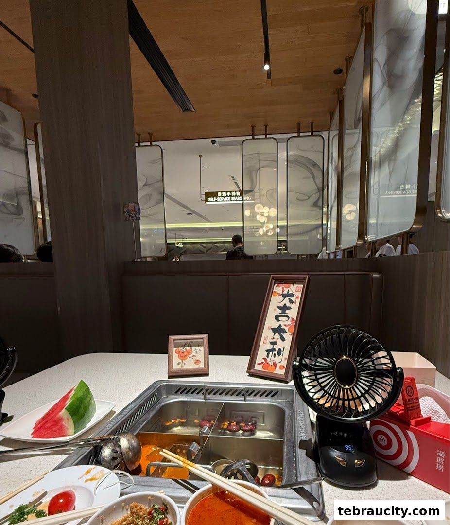 A table at Haidilao Hot Pot with a simmering pot, watermelon slices, and a small fan.