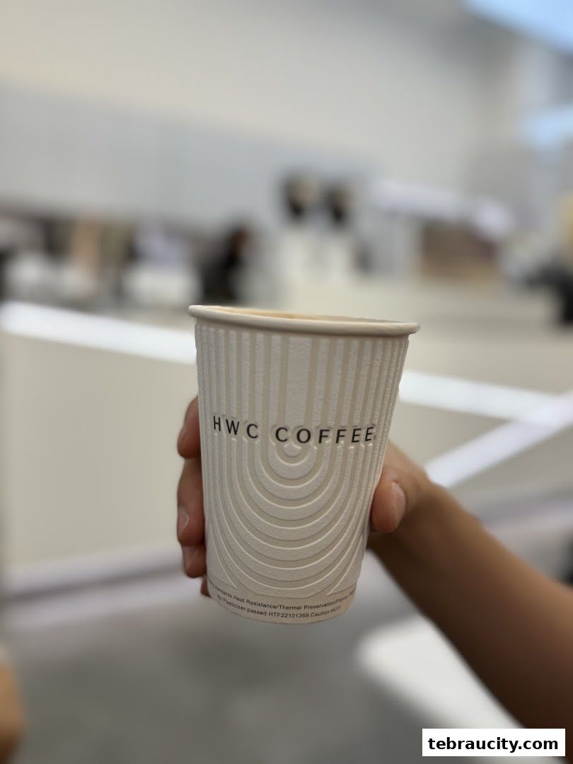 A hand holds a white paper cup with the HWC Coffee logo and a beverage inside.