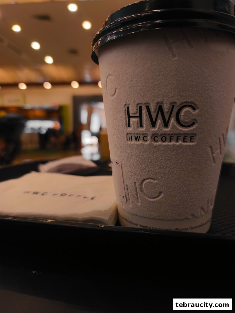 A close-up of a white HWC Coffee cup with a black lid and a stack of napkins.