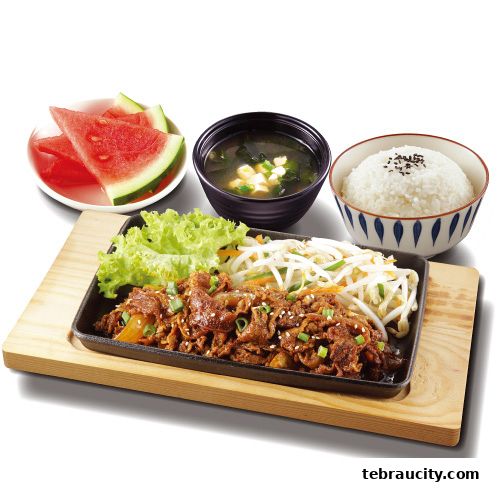 A sizzling hot plate of Korean BBQ beef with rice, soup, and fresh watermelon slices.