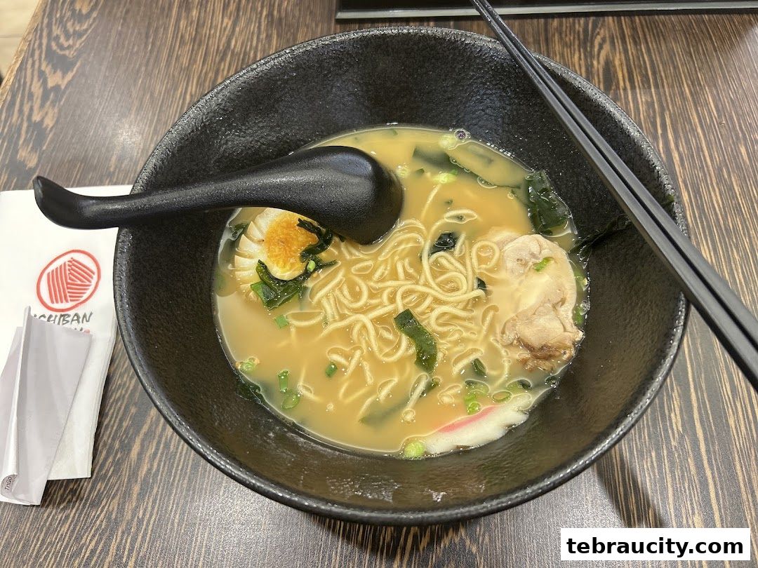 A steaming bowl of ramen with noodles, egg, meat, and seaweed, served with chopsticks and a spoon.