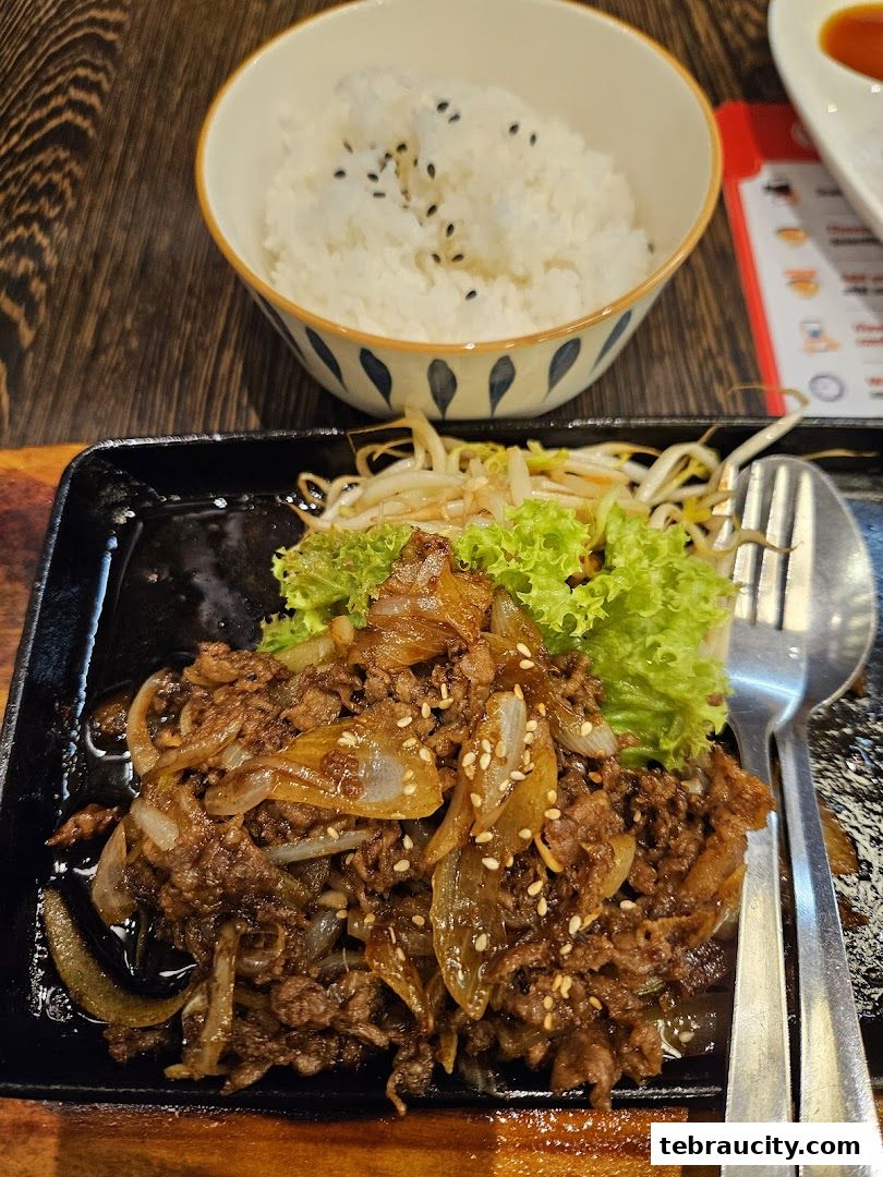 A sizzling plate of beef with onions and bean sprouts, served with a bowl of rice.