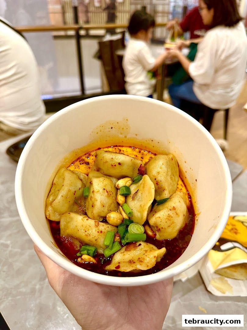 A hand holds a bowl of dumplings in chili oil, garnished with peanuts and scallions.