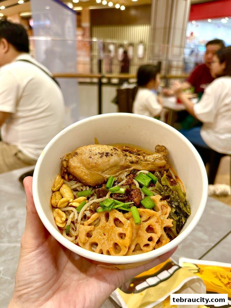 A hand holds a bowl of noodles with chicken, lotus root, peanuts, and vegetables.