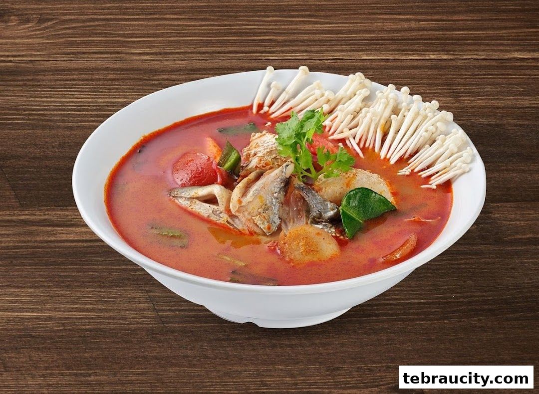 A bowl of Tom Yum soup with fish, mushrooms, and herbs on a wooden table.