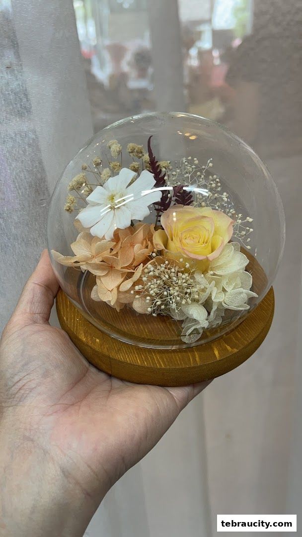 A hand holds a glass dome filled with preserved flowers, including a yellow rose.