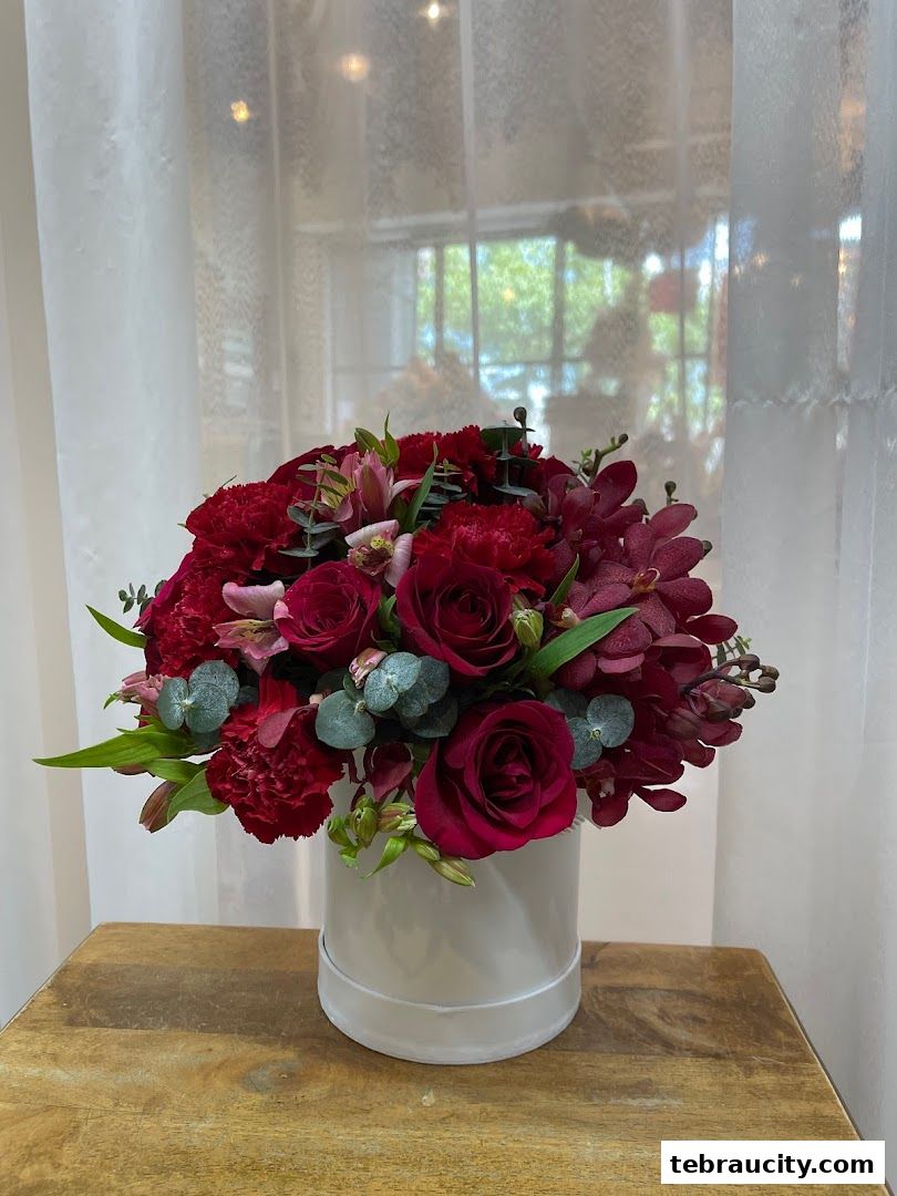 A luxurious arrangement of red roses, carnations, and orchids in a white hat box.