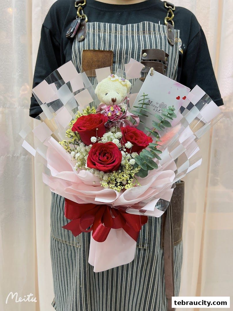 A person in an apron holds a bouquet of red roses with a teddy bear.