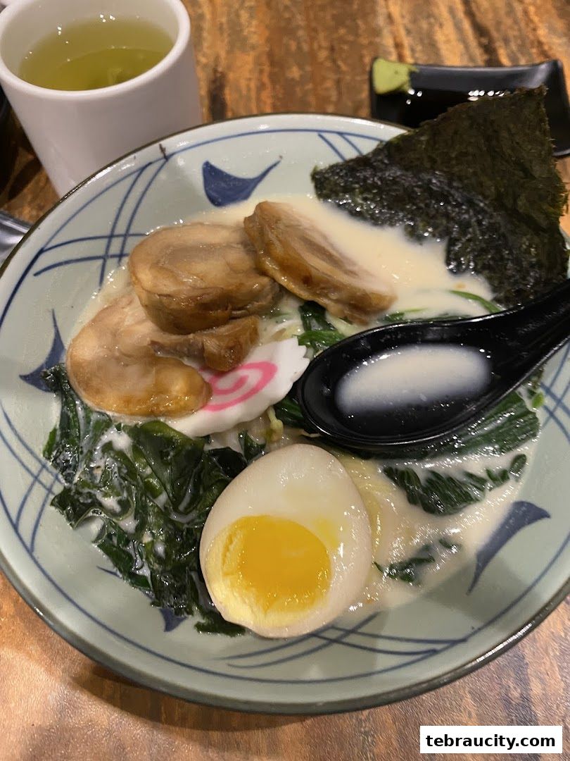 A steaming bowl of ramen with pork, egg, seaweed, and greens, served with green tea.