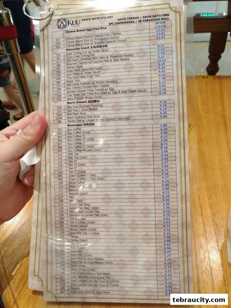A person holds a laminated menu from Kuu By Tea Garden, listing various food and beverage items with prices.