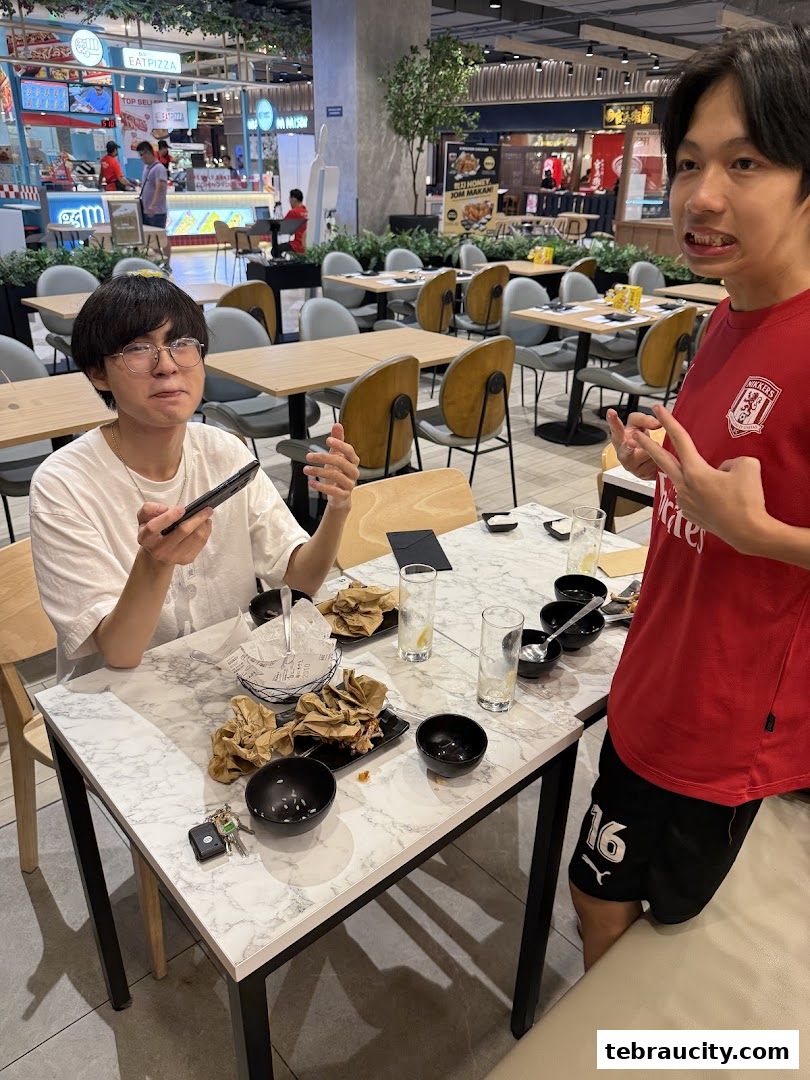 Two young men are sitting at a table in a food court, with food remnants on the table.
