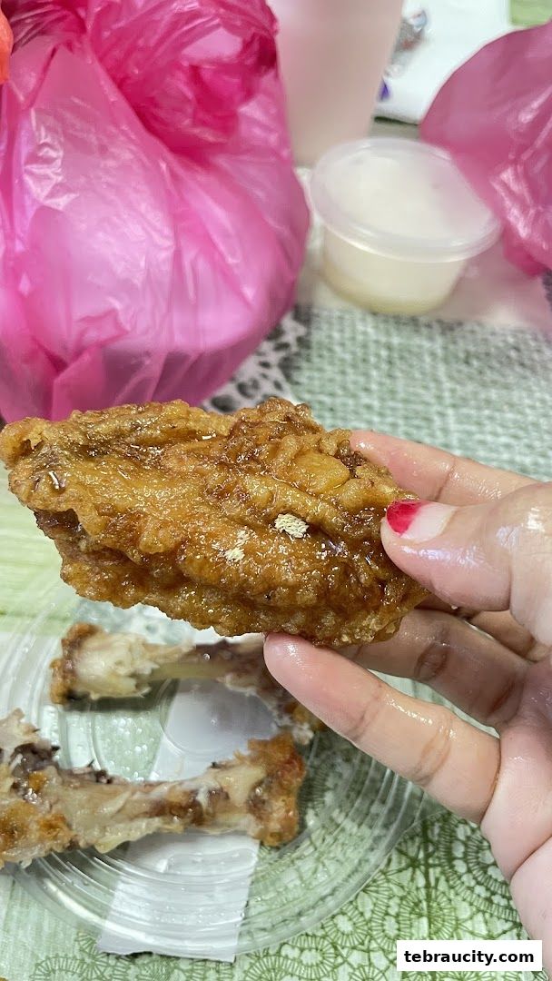 A hand holding a crispy fried chicken wing with a side of chicken bones and pink plastic bags.