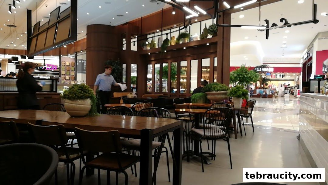 Interior view of a cafe with wooden tables, chairs, and plants, showing mall patrons in the background.