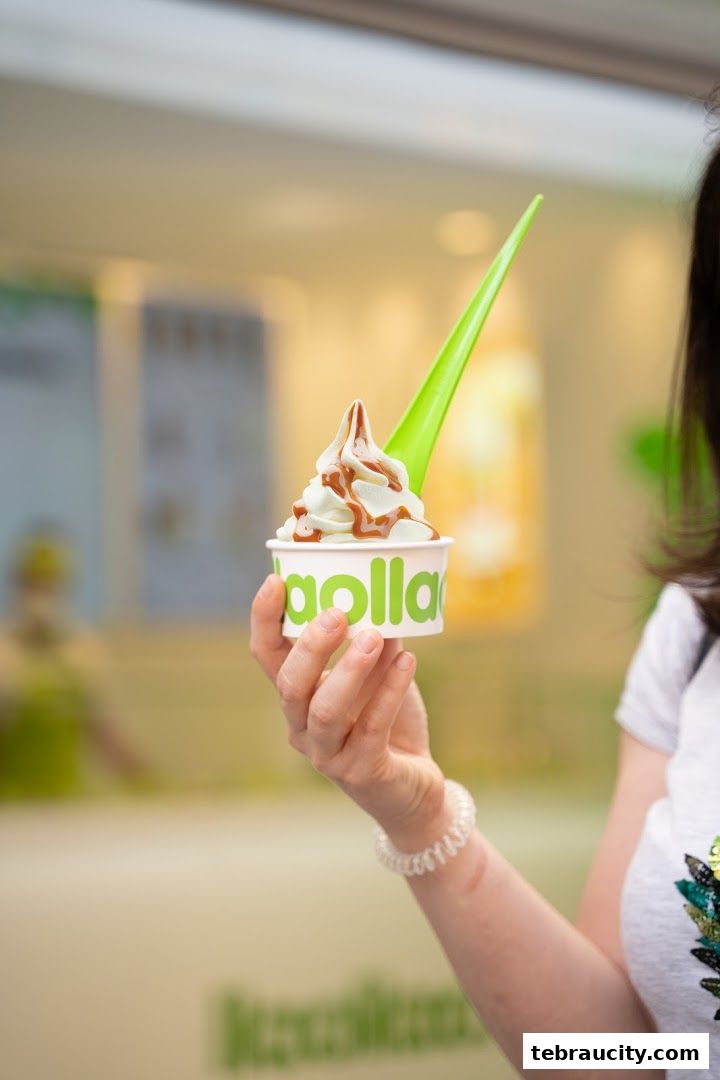 A hand holds a cup of frozen yogurt with caramel drizzle and a green spoon.