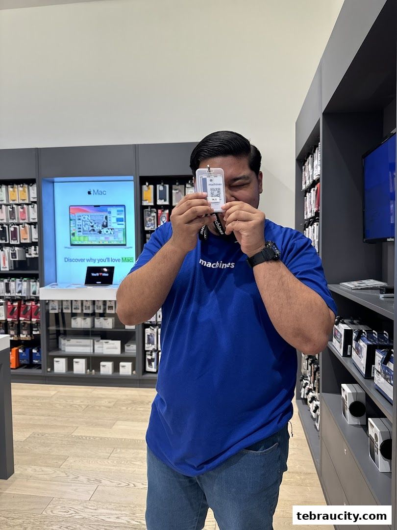 A man in a blue Machines t-shirt holds up an ID badge in an Apple Reseller Store.