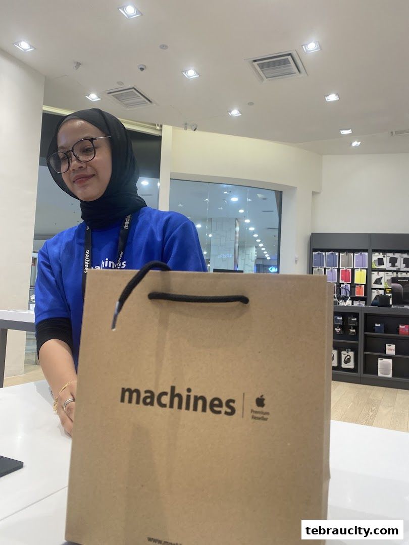 A smiling employee stands behind a counter with a Machines Apple Premium Reseller shopping bag.