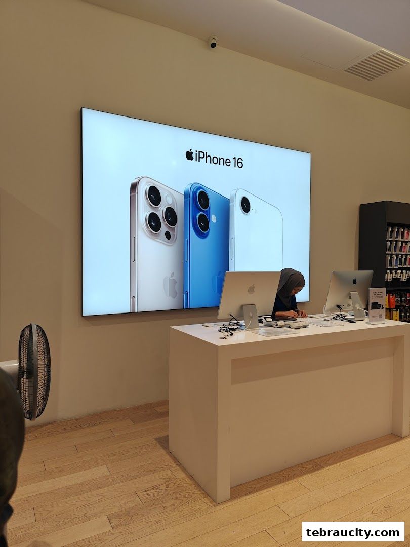 An Apple Reseller Store counter with iPhones displayed on a large screen and a staff member working.