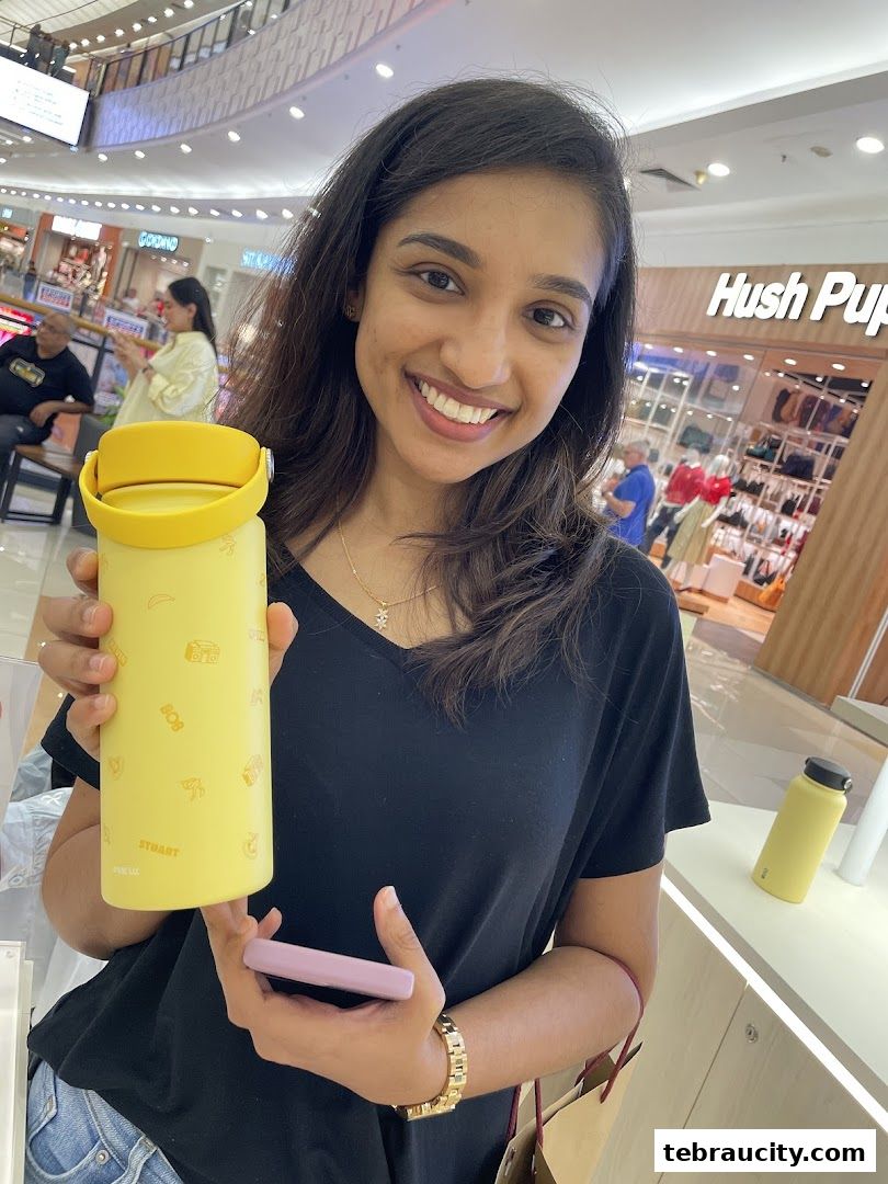 A smiling woman holds a yellow water bottle in a shopping mall.
