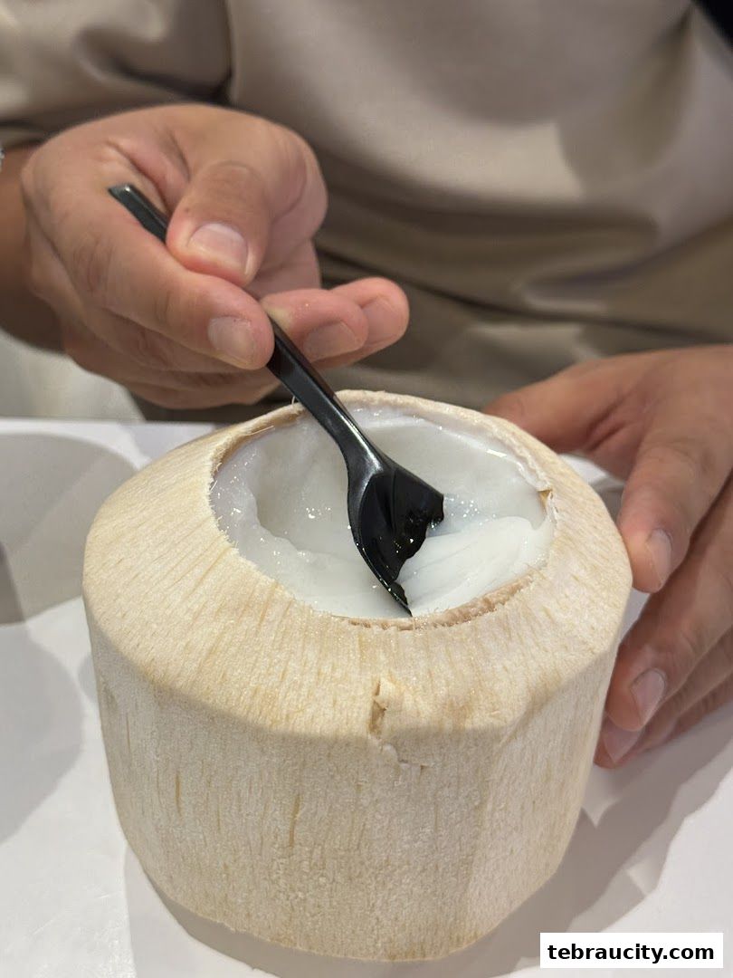 A person is scooping fresh coconut flesh from a whole coconut with a black spoon.