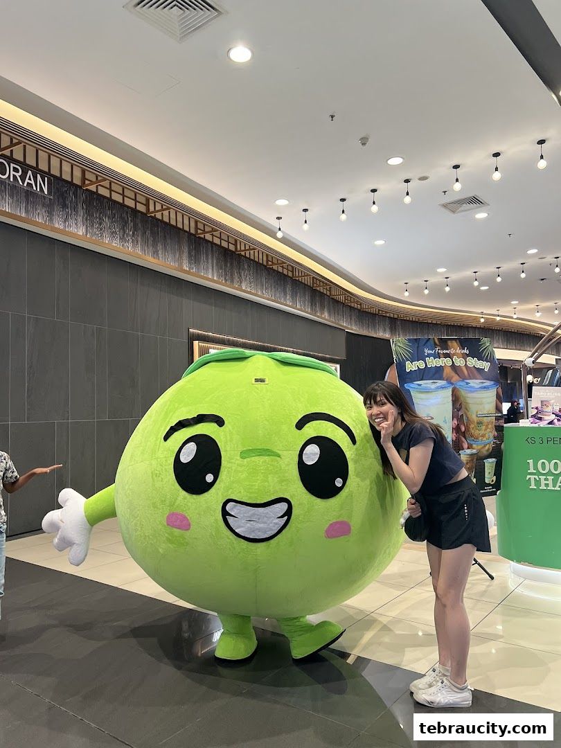 A woman poses with a large, smiling coconut mascot in a shopping mall.