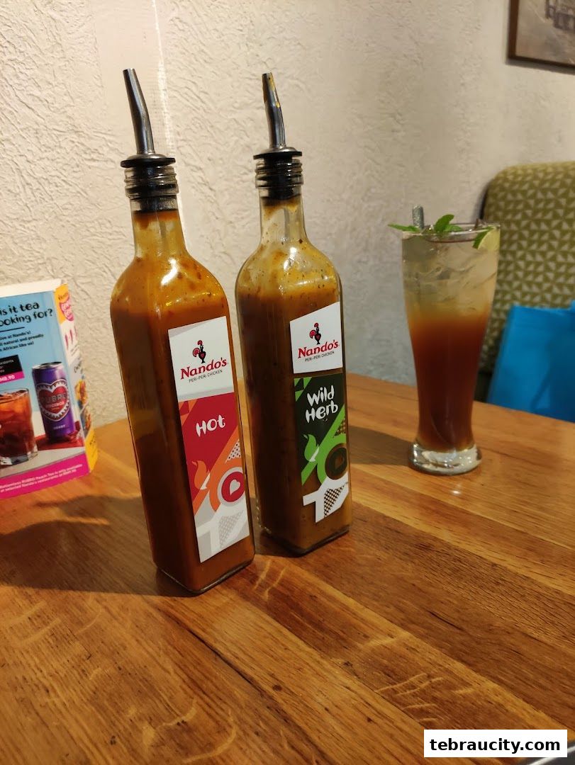Two bottles of Nando's peri-peri sauce and a refreshing drink on a wooden table.