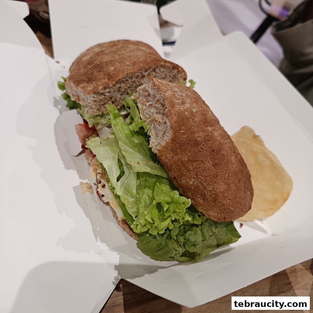 A hearty sandwich with fresh lettuce and a side of chips in a white takeaway box.
