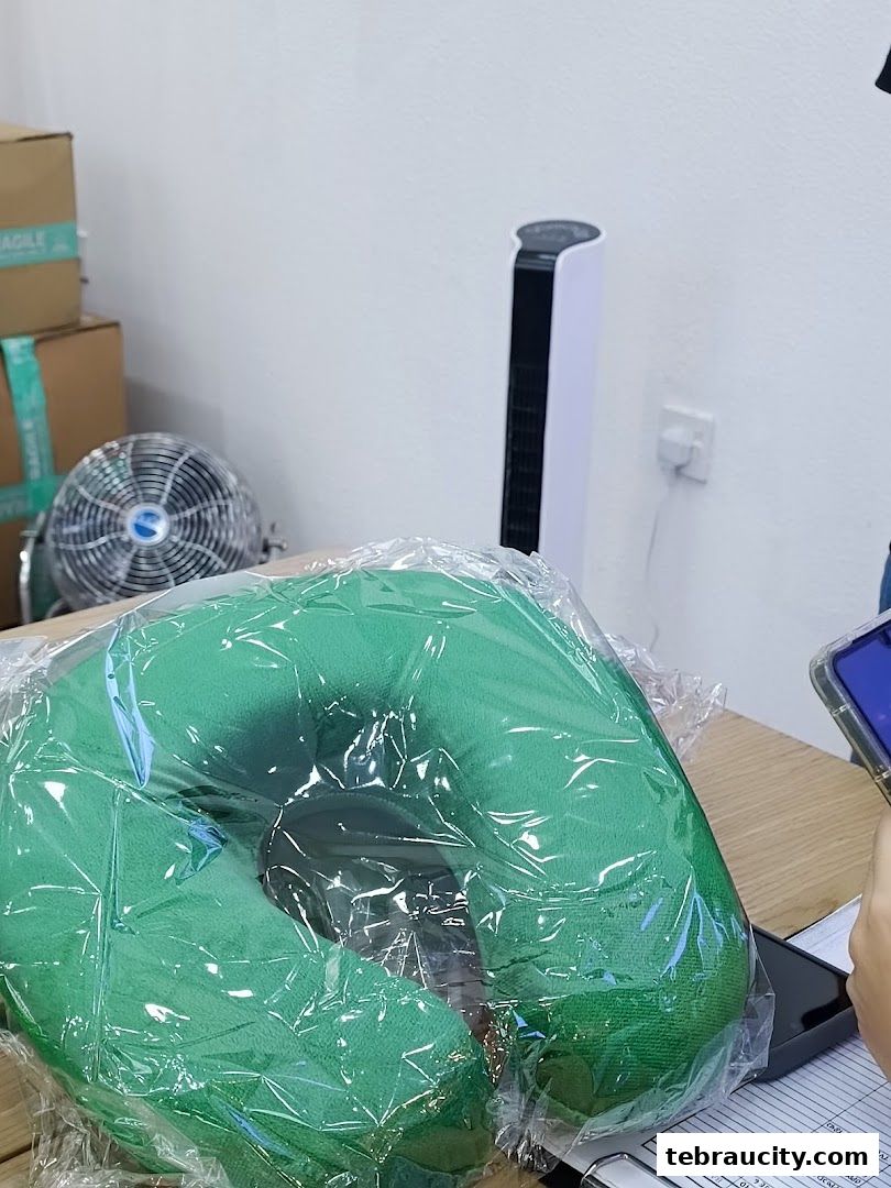 A green neck pillow is displayed on a table with boxes and fans in the background.