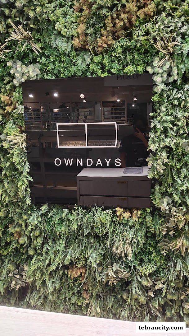 The OWNDAYS shop front features a modern design with a lush green plant wall and a prominent logo.