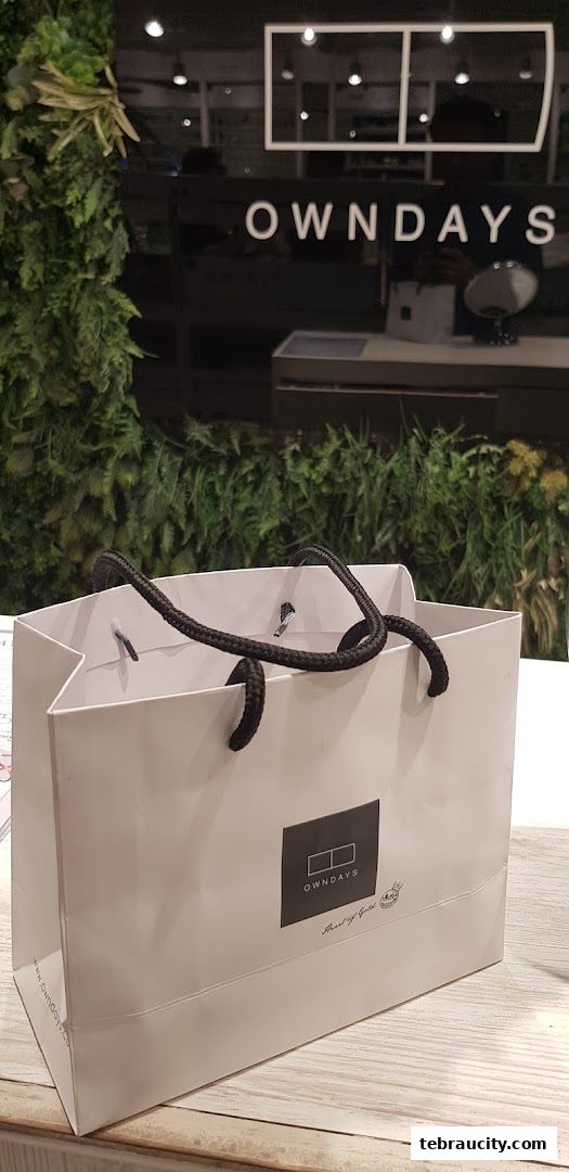 A white shopping bag with the OWNDAYS logo and black handles sits on a wooden surface.