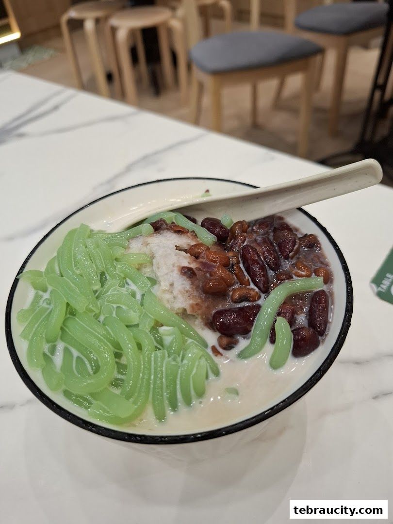 A close-up of a bowl of Chendol dessert with green jelly noodles and red beans.