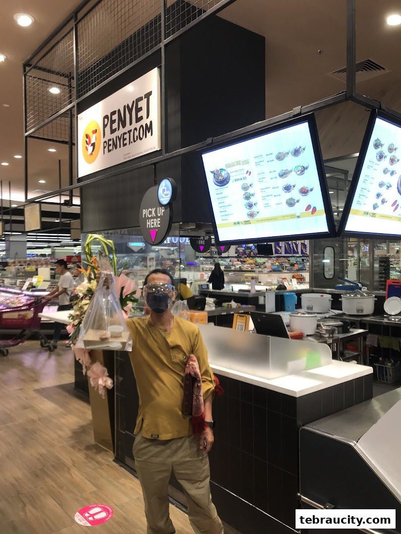 A man holds a takeaway food bag in front of the PenyetPenyet counter with digital menus displayed.